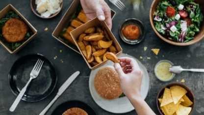 Hands sharing fries over table with burger, salad, chips and assorted dishes during meal.